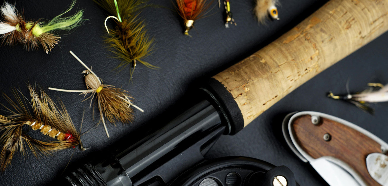 Flat lay of JP Fly Co-style fly fishing gear with detailed hand-tied flies, a cork rod handle, and reel arranged on a clean, dark background.