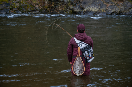 Angler wading in river casting fly line with landing net and pack, BC-style rocky river conditions
