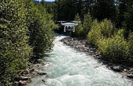A drone hovers above a fast-flowing river in British Columbia, surrounded by dense forest, capturing an aerial view of the rugged natural landscape.