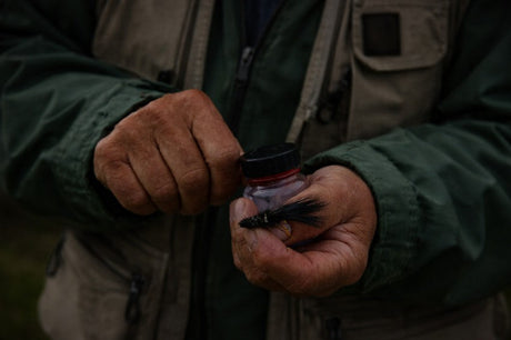 Close-up of an angler’s hands holding a black Wooly Bugger fly from JP Fly Co, with a small container, set against outdoor fishing gear.