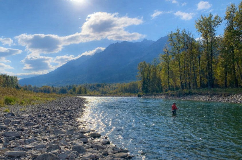 A lone angler stands quietly in a flowing river, surrounded by forest and mountains, reflecting a calm and mindful moment in nature.