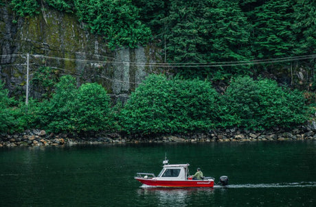 Small red motorboat traveling along calm coastal water with a forested rocky shoreline in British Columbia, featuring an angler in outdoor gear navigating the vessel